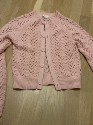 Rosa kofta från Gina Tricot - Söt ljusrosa kofta från Gina Tricot med stickat hålmönster och stora knappar framtill. Koftan har ribbade muddar vid ärmslut och nederkant, samt rund halsringning. Perfekt för lager-på-lager och en gullig look.