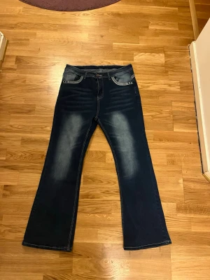 Mörkblå bootcut jeans med slitningar - Säljer ett par mörkblå bootcut jeans med snygga slitningar framtill och dekorativa bakfickor med nitar. Jeansen har klassisk femficksmodell och kontrastsömmar. Perfekta för dig som gillar en retroinspirerad look och vill ha ett par jeans med lite vidare ben.