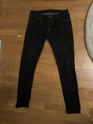 Mörkblå  jeans från Nudie - Säljer ett par mörkblå  jeans från Nudie med klassiska kontrastsömmar och femficksmodell. Jeansen har en smal passform och är tillverkade i stretchigt denimtyg för extra komfort. Perfekta för dig som gillar en clean och stilren look. W31 L34