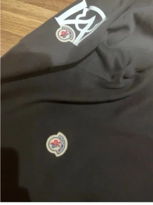 Svart sweatshirt från Moncler - Svart sweatshirt från Moncler med klassisk rund hals och Moncler-logga på bröstet. Ärmen har en stor vit grafisk detalj och logga. Tröjan har ribbade muddar vid ärmslut och nederkant. Perfekt för en stilren och trendig look.