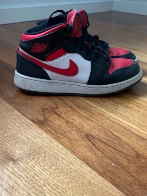 Nike Air Jordan 1 Mid röd/svart/vit - Säljer ett par Nike Air Jordan 1 Mid sneakers i klassisk röd, svart och vit färgkombination. Skorna har svart läder runt tån och snörningen, röd tåbox och häl, samt vit sidopanel med röd swoosh. Tjock vit sula och svart yttersula. Perfekta för dig som gillar streetwear.