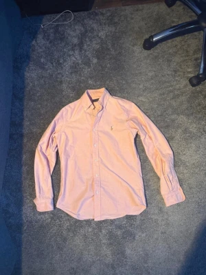 Ljusrosa skjorta från Ralph Lauren - Klassisk ljusrosa skjorta från Ralph Lauren med button-down krage och broderad logga på bröstet. Skjortan har långa ärmar och vita knappar framtill. Perfekt till både jeans och chinos för en clean look. Passar storlekar s/M