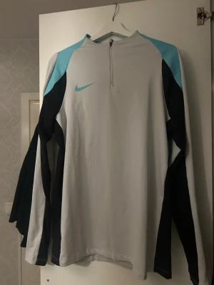 Nike långärmad träningströja - Nike långärmad träningströja i ljusgrått med svarta och turkosa detaljer. Tröjan har halv dragkedja framtill och är tillverkad i ett lätt, funktionsmaterial som passar perfekt för träning. Snygg sportig design med Nike-logga på bröstet.
