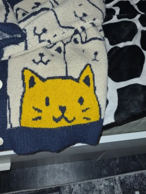 Beige kofta med kattmotiv från Aelfric Eden - Unik beige kofta med stora kattmotiv i svart och en gul katt framtill. Koftan har djupblå ribbade muddar, knäppning med vita knappar och V-ringad hals. Perfekt för dig som gillar statement-plagg och gulliga detaljer.