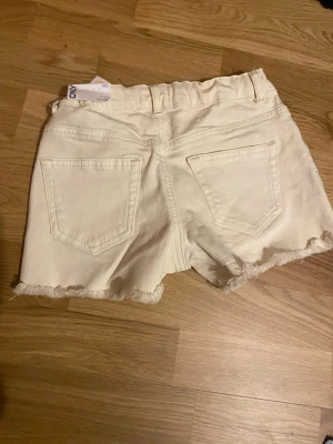 Vita jeansshorts från ONLY - Säljer ett par vita jeansshorts från ONLY med råa kanter och klassisk femficksdesign. Shortsen har dragkedja och knapp framtill samt bälteshällor. Perfekta för varma dagar och ger en avslappnad look. Har testat dem en gång och de passade inte så de är helt i nytt skick!