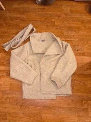 Beige teddyjacka med bälte - Mysig ljusbeige teddyjacka med bred krage och tillhörande bälte. Jackan har tryckknappar framtill som man stänger med. 