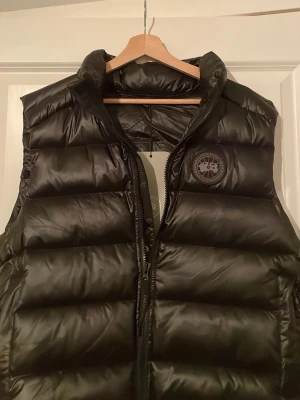 Svart dunväst från Canada Goose - Snygg svart dunväst från Canada Goose med hög krage och diskret logotyp på bröstet. Västen har en glansig finish och breda, vadderade paneler som ger en modern look. Perfekt för lager-på-lager och håller dig varm under kyliga dagar.