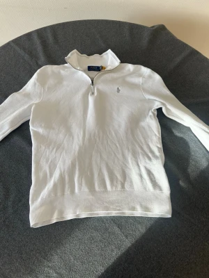 Vit halvzip från Polo Ralph Lauren - Klassisk vit tröja från Polo Ralph Lauren med halvzip och ståkrage. Tröjan har diskret broderad logga på bröstet och är tillverkad i ett mjukt, stickat material. Perfekt för en clean och stilren look. Storlek M men passar S.