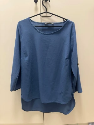 Blå topp med trekvartsärm - En enkel blå blus med rund halsringning och trekvartsärm. Toppen har en lös passform och något längre bakstycke för en avslappnad look. Perfekt att matcha med jeans eller kjol.