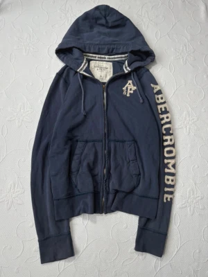 Mörkblå hoodie från Abercrombie - Mörkblå hoodie med dragkedja från Abercrombie & Fitch. Stor logga i vitt längs ena ärmen och ett broderat A&F-märke på bröstet. Tyvärr finns två små hål på ärmen samt att det högra snöret är sliten.