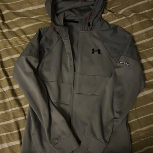 Grå träningsset från Under Armour - Säljer ett grått träningsset från Under Armour med hoodie och matchande byxor. Hoodien har dragkedja, huva och logga på bröstet. Byxorna har resår i midjan, dragkedjeficka och logga på benet. Perfekt för gymmet eller löprundan.