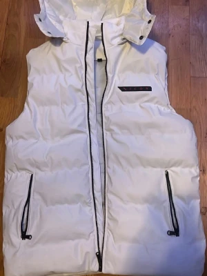 Vit dunväst med huva från ICON - Snygg vit dunväst från ICON med avtagbar huva och svarta dragkedjor. Västen har två fickor med dragkedja och en logga på bröstet. Perfekt för lager på lager och streetwear-stil.