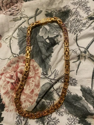 Guldfärgat kedjehalsband med stenar - Snyggt halsband i guldfärg med kraftig kedjedesign och dekorativt spänne prytt med små gnistrande stenar. Halsbandet har en unik, flätad look och känns riktigt statement. Perfekt för dig som vill sticka ut med accessoarerna.