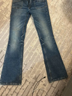Blå bootcut jeans med slitningar - Säljer ett par blå bootcut jeans med snygga slitningar och ljusare partier framtill. Jeansen har klassisk femficksdesign och lågmidja. Perfekta för dig som gillar en retroinspirerad look med utsvängda ben.
