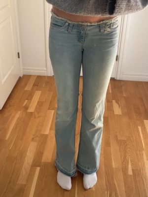 Ljusblå bootcut jeans - Säljer ett par ljusblå bootcut jeans med klassisk femficksmodell och låg midja. Jeansen har slitna detaljer vid benslut och är tillverkade i mjukt denimtyg. Perfekta för en avslappnad och trendig look.