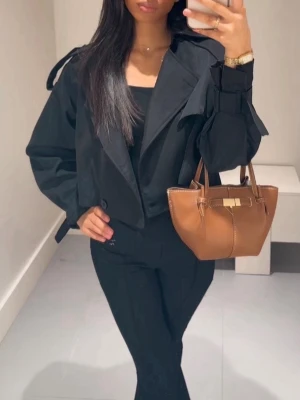 Kort svart trenchcoat  - Snygg svart cropped trenchcoat från  med bred krage, dubbelknäppning och trendiga axel- och ärmdetaljer. Jackan är knappast använd och i nyskick. 