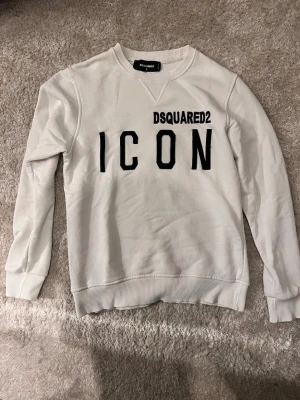 Vit sweatshirt från Dsquared2 ICON - Säljer en vit sweatshirt från Dsquared2 med svart text 'ICON' och logga på bröstet. Tröjan har rund halsringning, ribbade muddar och är tillverkad i mjukt bomullsmaterial. Perfekt för en trendig och avslappnad stil.