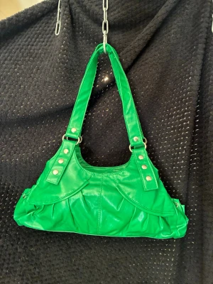 Grön axelväska med nitar - Cool grön handväska i syntetmaterial med breda axelband och silverfärgade nitar. Väskan har en unik form, dekorativa sömmar och dragkedjeficka på baksidan. Perfekt för dig som vill sticka ut med en färgglad accessoar.
