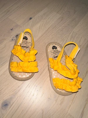 Gula sandaler med volang  - Säljer ett par söta gula sandaler från Myrapp med dubbla volangband och öppen tå. Sandalerna har elastiska remmar för enkel påtagning och en beige sula med små blommor. Perfekta för sommarens alla äventyr!