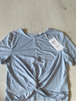 Ljusblå twistad topp från VILA - Snygg ljusblå topp från VILA med twistad detalj framtill. T-shirten har korta ärmar och är gjord i mjuk bomull, perfekt för dig som gillar en enkel men trendig look. Rund halsringning och stilren design som är lätt att matcha.