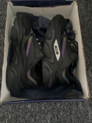 Svarta Dior sneakers med meshdetaljer - Säljer ett par svarta Dior sneakers med chunky sula och meshpaneler för extra ventilation. Skorna har diskreta regnbågsskimrande detaljer på sidan och klassisk snörning. Märkeslogga på sidan och bak. Perfekta för dig som gillar streetwear och exklusiv stil.