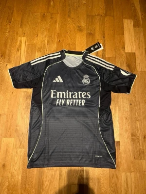 Real Madrid away kit 25/26 X Mbappé 10 - Snygg svart Real Madrid matchtröja från Adidas med vita detaljer och tryck. Framsidan har klubbmärke och Emirates Fly Better-logga, baksidan har Mbappé och nummer 10. Tillverkad i lätt och ventilerande material, perfekt för fotbollsträning eller match.
