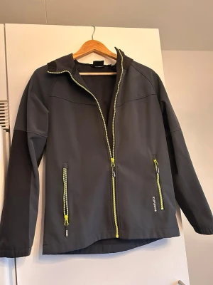 Svart vindjacka från Icepeak - Snygg svart vindjacka från Icepeak med luva och dragkedjor i neonfärg. Jackan har två framfickor med dragkedja och elastiska muddar vid ärmsluten. Materialet är slätt och lätt, perfekt för blåsiga dagar. Reflektiv detalj baktill för extra synlighet. Storlek 164. 