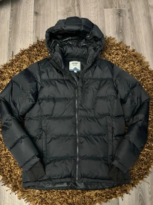 Svart dunjacka från Tuxer M - Snygg svart dunjacka från Tuxer med huva och dragkedja framtill. Jackan har två sidofickor med dragkedja och diskret logga på bröstet. Perfekt för kalla dagar och enkel att matcha med olika outfits.