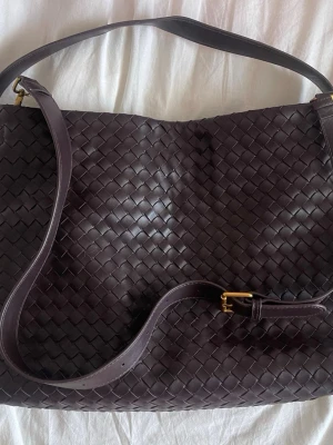 Mörkbrun flätad axelväska i läder - Snygg mörkbrun axelväska i flätat läder med justerbar axelrem och guldfärgade metalldetaljer. Väskan har en mjuk, rektangulär form och klassisk design som passar till många olika stilar. Perfekt för dig som gillar tidlösa accessoarer.