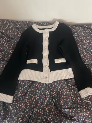 Svart kofta med vita detaljer - Snygg svart kofta med vita ribbstickade kanter och pärlemorknappar. Koftan har två dekorativa fickor framtill och en croppad passform. Perfekt för dig som gillar klassisk men trendig stil. Det e storlek M och den är från New Yorker.