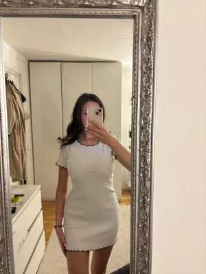 Vit ribbad kortklänning med svarta kanter - Säljer en vit ribbad kortklänning med svarta vågiga kanter vid ärmslut och nederkant. Klänningen har en figurnära passform och korta ärmar. Perfekt för dig som gillar enkel men snygg stil.