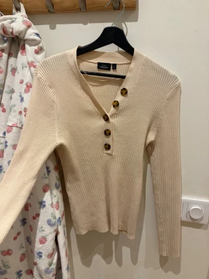 Beige ribbstickad tröja KappAhl - Snygg beige ribbstickad tröja från KappAhl med v-ringning och fyra stora, mönstrade knappar framtill. Tröjan har lång ärm och är gjord i ett mjukt, stretchigt material som sitter skönt på kroppen. Perfekt till jeans eller kjol.