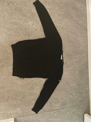 Svart stickad tröja från Stone Island - Säljer en svart stickad tröja från Stone Island med klassisk rund hals och den ikoniska loggan på vänster ärm. Tröjan har en enkel, stilren design och är ribbstickad vid ärmslut och nederkant. Perfekt för dig som gillar streetwear och snygga basplagg.