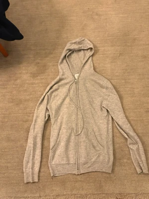 Ljusgrå zip-hoodie med huva - En stilren ljusgrå hoodie med dragkedja framtill och två fickor. Tröjan har ribbade muddar vid ärmslut och nederkant samt en klassisk huva med dragsko. Perfekt för en avslappnad och enkel look.
