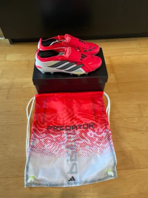 Adidas Predator röda fotbollsskor - Säljer ett par Adidas Predator fotbollsskor i en grymt snygg röd färg med svarta och vita detaljer. Skorna har klassisk låg profil, snörningslös design och mönstrad ovandel för bättre bollkontroll. Kommer med matchande Predator-påse.
