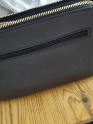 Svart axelväska med gulddragkedjor - Snygg svart axelväska i slätt material med två stora fack och guldfärgade dragkedjor. Väskan har en justerbar axelrem och en praktisk ytterficka med dragkedja. Perfekt för att bära det viktigaste och ger en stilren look.