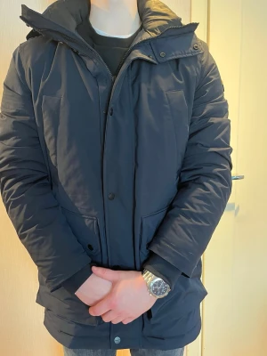 Marinblå J.Lindeberg vinterjacka med huva - Snygg marinblå vinterjacka med huva och stora framfickor. Jackan har dragkedja och knappslå framtill samt justerbar huva. Perfekt för kalla dagar och har en klassisk, rak passform. Fodrad insida för extra värme.