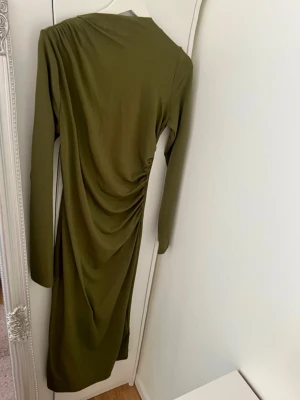 Olivgrön långärmad klänning H&M - Snygg olivgrön långärmad klänning från H&M med draperad detalj på sidan och asymmetrisk axel. Klänningen har en smickrande passform och är tillverkad i ett mjukt syntetmaterial som känns bekvämt mot huden.