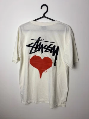 Vit Stüssy t-shirt med rött hjärta - Vit t-shirt från Stüssy med stort svart logotryck och ett rött hjärta på ryggen samt mindre logga och hjärta på bröstet. Klassisk passform och rund halsringning. Perfekt för dig som gillar streetwear och vill sticka ut.