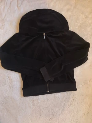 Svart velourhoodie Juicy Couture - Snygg svart hoodie i mjuk velour från Juicy Couture. Klassisk ziptröja med huva och ribbade muddar. Perfekt för en chill och trendig look. Dragkedja framtill och diskret logga på insidan. Superbekväm och enkel att matcha.