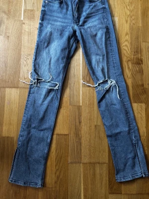 Gråa slitna raka jeans med hål - Säljer ett par blå jeans med raka ben och stora slitna hål över knäna. Jeansen har klassisk femficksdesign och är i en tvättad denim med råa kanter vid hålen. Perfekta för en avslappnad och trendig streetstil.