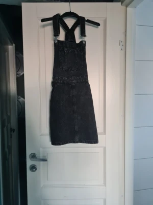 Svart denimskort med hängslen - Säljer en svart jeansskort med cool tvätt och justerbara hängslen. Skorten har en ficka framtill på bröstet och två bakfickor. Rak passform och klassisk jeanslook som passar perfekt till en avslappnad stil.