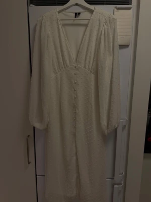 Vit långklänning med prickigt mönster - Elegant vit långklänning med diskret prickigt mönster, v-ringning och långa ärmar. Klänningen har knappar framtill och är tillverkad i ett lätt, luftigt material som ger en romantisk känsla.