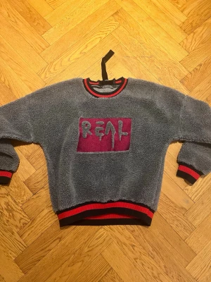 Grå teddytröja med röd REAL-tryck - Mysig grå teddytröja med röd och svart ribbade muddar vid ärmslut, hals och midja. På bröstet finns ett stort rött fält med texten 'REAL' i grått. Perfekt för dig som gillar streetwear och vill ha något riktigt skönt och trendigt.