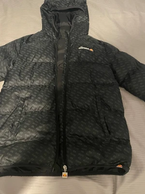 Svart dunjacka från Ellesse - Snygg svart pufferjacka från Ellesse med heltryckt logomönster och huva. Jackan har dragkedja framtill, två sidofickor och Ellesse-logga broderad på bröstet. Perfekt för kalla vinterdagar och riktigt trendig look. Hyfsat varm men fungerar att ha på hösten och våren med. Storleken: 152-158