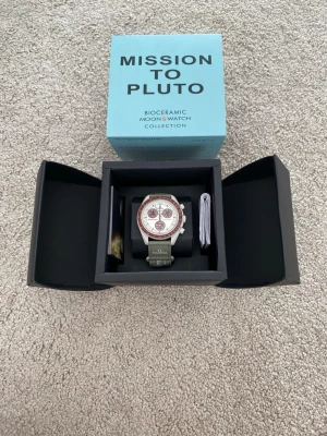 Omega x Swatch Mission to Pluto klocka - Säljer en Omega x Swatch Speedmaster MoonSwatch Mission to Pluto. Klockan har en beige urtavla med vinröda detaljer och tachymeter, samt ett grönt textilband med Omega-logga. Snygg och unik design från Bioceramic MoonSwatch Collection.