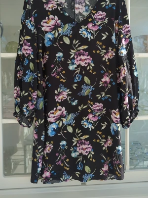 Blommig svart klänning från Xlnt - Svart klänning från Xlnt med färgglatt blommönster i rosa, blått, lila och grönt. Klänningen har v-ringning och trekvartsärmar med resår vid ärmslut. Perfekt för dig som gillar en feminin och färgstark stil.