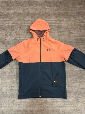 Tvåfärgad vindjacka från Under Armour - Snygg vindjacka från Under Armour i orange och mörkblått. Jackan har huva med dragsko, hel dragkedja framtill och två sidofickor. Tillverkad i lätt och tåligt material, perfekt för blåsiga dagar. Under Armour-logga på bröstet och justerbar nederkant.