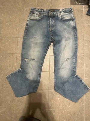 Blå slitna jeans med upprullade ben jack and Jones  - Säljer ett par blå jeans med slitningar och upprullade benslut. Jeansen har fem fickor, ljus tvätt och diskreta slitdetaljer på framsidan. Perfekta för en avslappnad och trendig look.