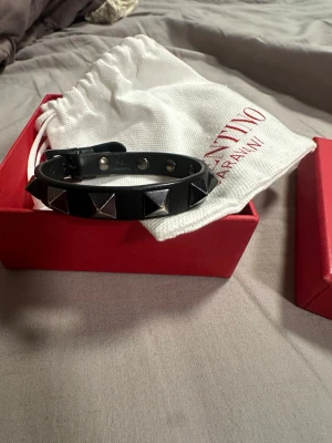 Svart Valentino armband med nitar - Snyggt svart armband från Valentino Garavani i äkta läder med silverfärgade pyramidformade nitar. Justerbart spänne och stilren design som ger en edgy vibe. Kommer med originalförpackning och dustbag och kvitto. På sista bilden kan ni se lite slitage ockdp 